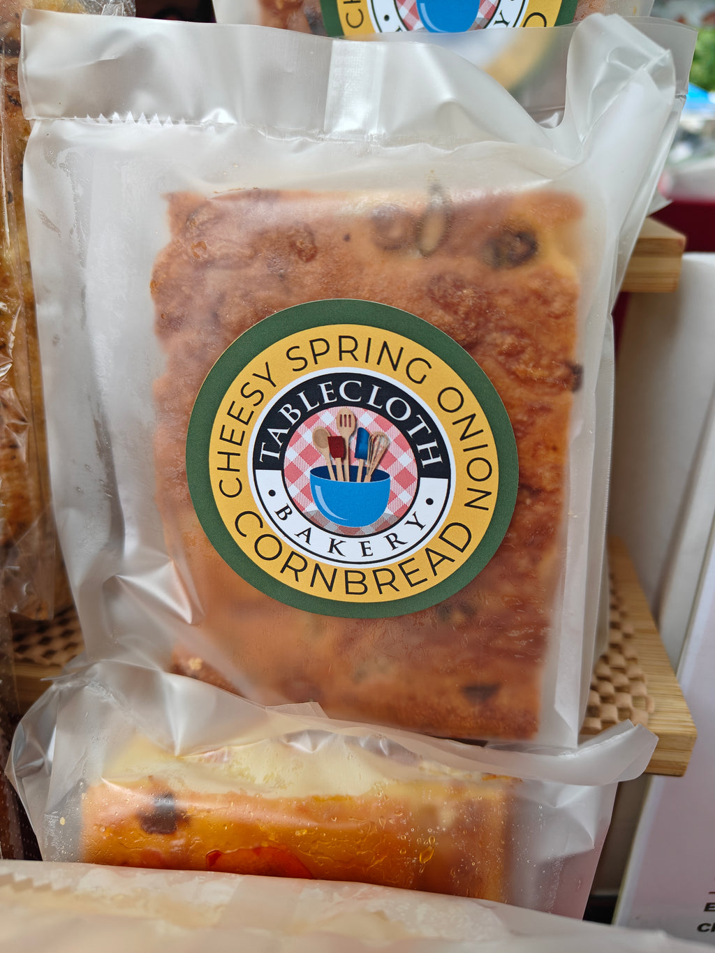 A packaged slice of cheesy spring onion cornbread from Tablecloth Bakery, sealed in a clear plastic bag with a prominent green and gold circular logo on the front.