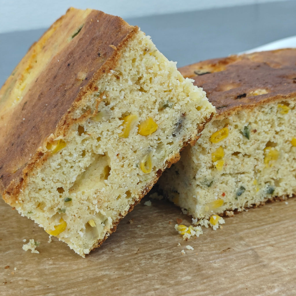 Two slices of a corn and vegetable bread leaning against each other on a wooden board, showing a textured crumb with visible corn kernels and green vegetable pieces.